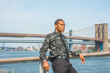 Fototapeta na wymiar African American businessman traveling in New York. Wearing black flower patterned shirt, tie, a young guy standing at harbor, thinking, waiting for you. Manhattan, Brooklyn bridges on background..
