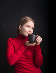 Beautiful young woman with cup of coffee, on black studio background. Smiling and cheerful woman holding a cup with hot tea. Winter girl drinking tea or coffee to warm up. Lifestyle studio photo.