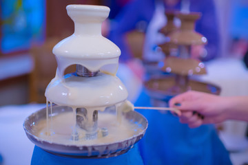 Woman holds a marshmallow into Chocolate fondue fountain with white chocolate. Sweets and dessert...