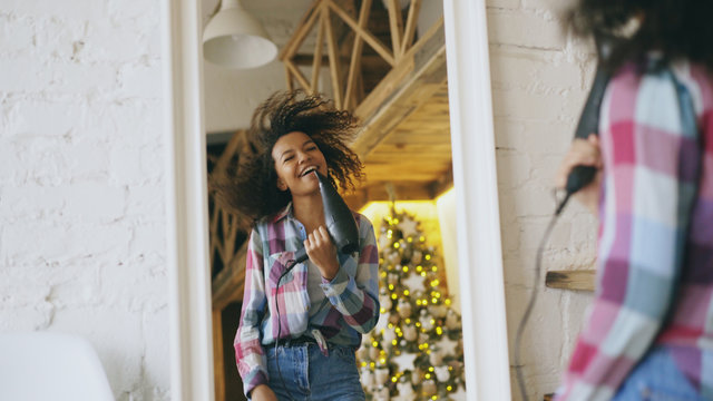 Curly Funny African American Girl Dancing And Singing With Hair Dryer In Front Of Mirror At Home