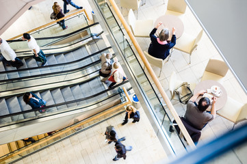 Typical scene in a shopping center : view from above of people of various sexes and ages taking the escalators, strolling along the walkways and having a snack on the patio of a cafe. - Powered by Adobe