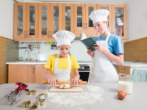 Little girl in the form of a chef with an apron and a cap blinks the dough with her hands, and next to her is her adult brother and reads a book on the background of the kitchen with a table 