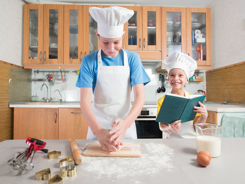 Schoolboy in the form of a cook in an apron and a cap holds a rolling pin in his hands and rolls out the dough, and next to it is his younger sister and reads a book