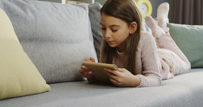 Lovely schoolgirl writing with a pencil in the notebook while lying on the grey sofa in the nice living room. Indoor