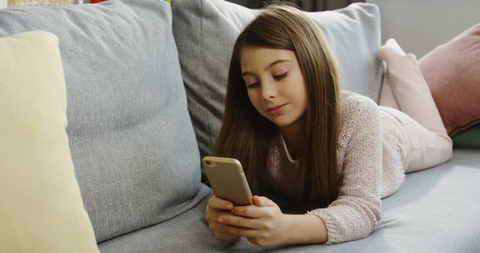 Pretty teen girl with long hair watching something on her smart phone while lying on the sofa with pillows at home on the weekend. Indoor