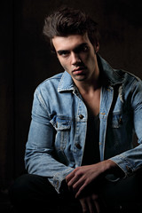 Handsome male model posing in fashion blue jeans jacket looking on dark shadow background. Closeup portrait