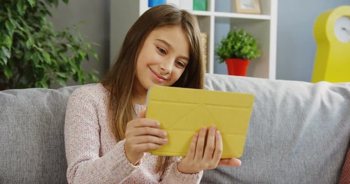 Pretty teenager girl using her tablet computer, scrolling and taping on it while sitting on the grey couch in the nice room. Insude