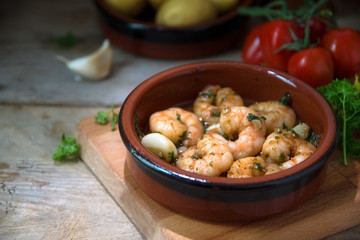 shrimps or prawns in garlic and olive oil in a tapas bowl, a spanish appetizer on a wooden board, potatoes, tomatoes and herbs in the background, dark rustic style