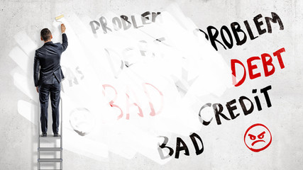 A businessman stands on a step ladder and paints over words Problem, Debt and Credit with a white paint roller. © gearstd