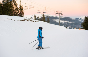 Fototapeta na wymiar Female skier on the middle of ski slope with ski lifts in background. Woman at ski resort wearing helmet, blue ski suit and goggles. Carpathian Mountains, Bukovel, Ukraine