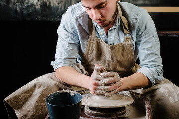 pottery, workshop, ceramics art concept - man working on potter's wheel with raw clay with hands, a male brunette sculpt a utensils near wooden table with tools, master in apron and a shirt