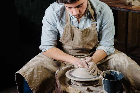 pottery, workshop, ceramics art concept - man working on potter's wheel with raw clay with hands, a male brunette sculpt a utensils near wooden table with tools, master in apron and a shirt