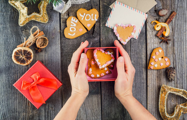 Top view female hands holding box with homemade cookies in form of hearts as gift for a loved one. Wooden table with greeting card, decor. Selective focus. Celebrating Valentine's Day. Festive gift.