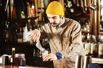 Portrait of hipster barman adding ice cubes to a cocktail at bar background.