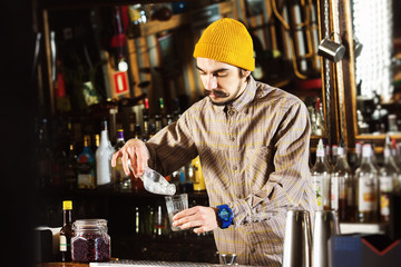 Portrait of hipster barman adding ice cubes to a cocktail at bar background.