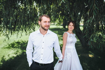 The bride and groom stand holding hands under a large tree.