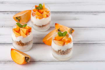 Persimmon creamy trifle in beautiful glasses, fresh ripe fruit slices on white wooden background. Healthy vegetarian food. Delicious dessert. Close up photography. Selective focus.