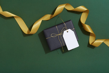 Black gift with blank gift tag and gold ribbon on dark green background, masculine, men, flat lay, Christmas
