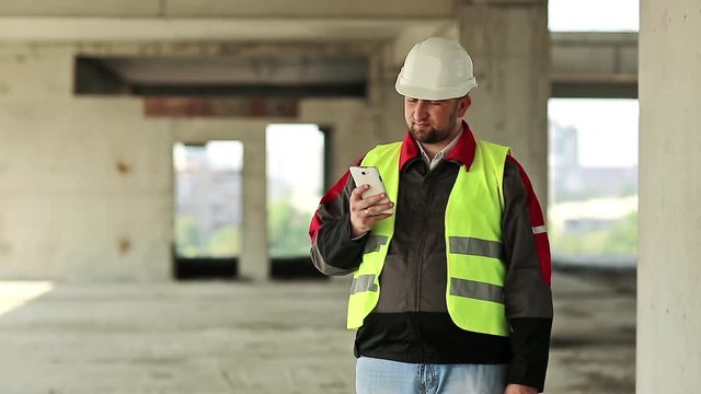 Workman in white helmet with smartphone at construction site. Builder in green vest with cell phone on project site. Worker holds in hands briefcase and mobile phone