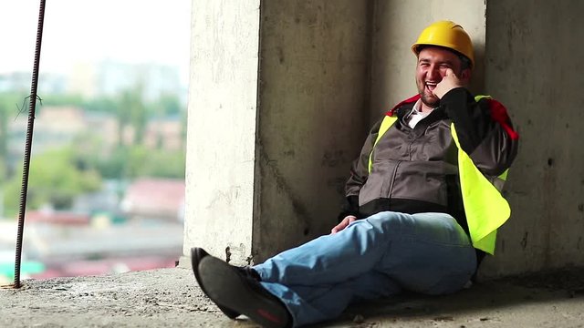 Cheerful worker sits on construction site. Funny builder in yellow hard hat sits on concrete floor, talks gesticulate and laughs