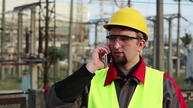 Service engineer at power plant speaks on smartphone. Worker talks on cell phone at heat electric power station near outdoor switchgear. Powerman in yellow hard hat communicates via mobile phone