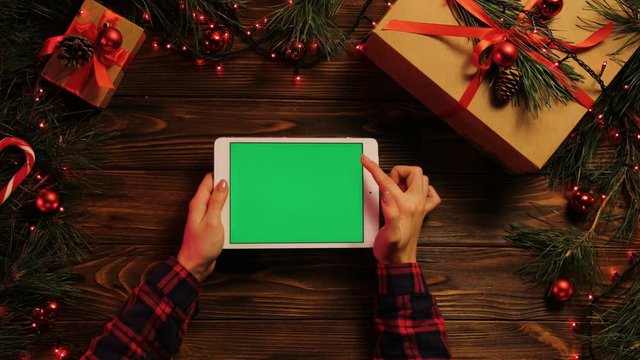 Top view of woman hands tapping, scrolling and zooming on tablet divice horizontally. The Christmas decorated brown desk with Christmas lights. Green screen, chroma key