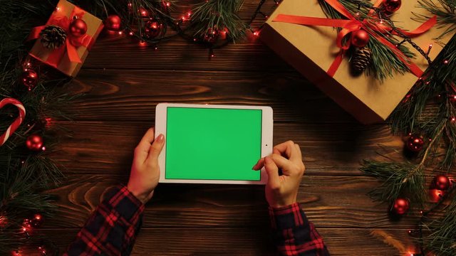 Top view. Female hands tapping, scrolling and zooming on tablet computer horizontally. The decorated wooden desk with Christmas lights. Green screen, chroma key.
