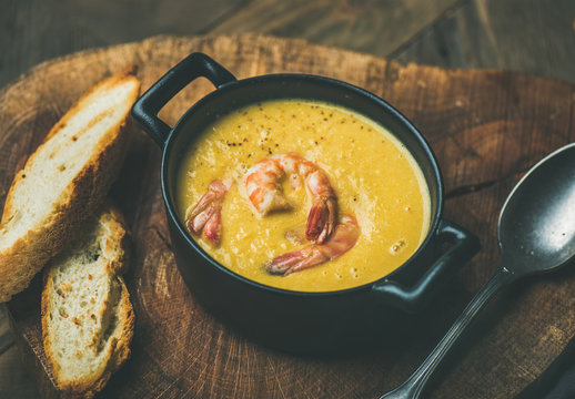 Corn creamy soup with shrimps served in individual pot with bread on board over rustic dinner table. Slow food, winter warming food concept