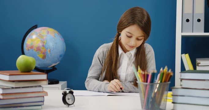 Portrait shot of the cute schoolgirl in glasses doing an exersice from a homework and writing in her copybook with a black pen at the table in the nice comfortable room with school stuff. Inside