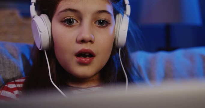 Close up of the pretty girl in big white headphones watching something scarry on the laptop and than smiling. Evening at home. Portrait. Inside