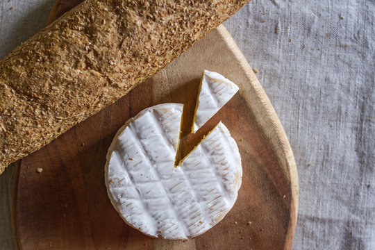 The Swiss Camembert formed cheese and a triangular piece of cheese on a wooden textured board and a grain baguette on a woven canvas background.