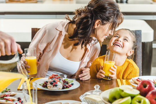 cheerful mother with daughter sitting at table with fresh juice and pancakes with berries at kitchen