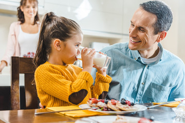 happy family at kitchen, daughter with father sitting at table with pancakes and looking at each other when daughter drinking milk