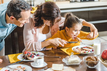 cheerful family together eating pancakes with berries at table, mother feeding daughter