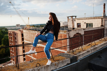 Portrait of a gorgeous young woman in black leather jacket, jeans and sneakers sitting on handrails on the roof with picturesque view of a park.