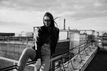 Obraz premium Portrait of a gorgeous young woman in black leather jacket, jeans and sneakers sitting on handrails on the roof with picturesque view of a park. Black and white photo.
