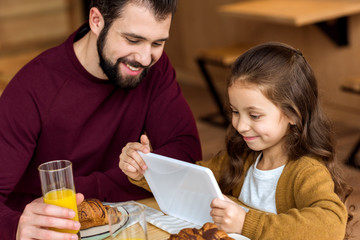 happy daughter looking at tablet while sitting with father in cafe