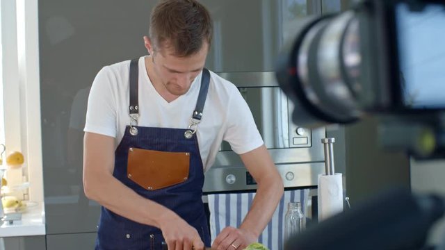 Handheld shot of concentrated male chef in apron cutting spring onion during shooting of cooking show