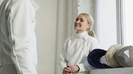 Two young smiling fencers man and woman sharing experience during break of fencing match indoors