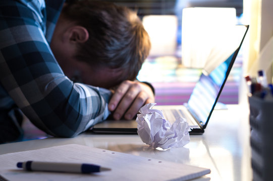 Sad And Tired Man In Home Or Modern Business Office At Night. Unemployed Job Seeker Or Businessman With Ripped Paper And Laptop On Table. No Motivation Or Inspiration. Writer's Block Or Out Of Ideas.