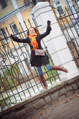 Girl in the red scarf and black coat jumps near the lattice fence