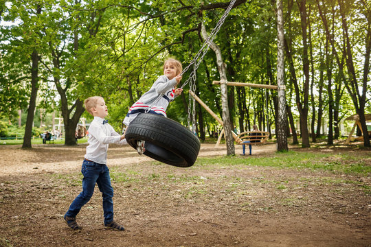 The brother rolls the younger sister on a tire swing. Children playing outdoors in summer.