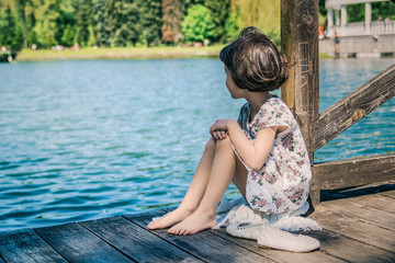 A little young girl is sitting on a wooden pier. Summer day