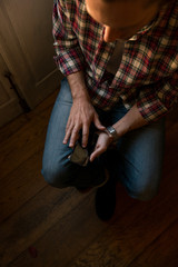 Young adult caucasian man with blonde ponytail wearing red squared shirt and blue jeans tap on black screen reflection smartphone near window in cozy home.