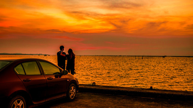 Silhouette of happiness couple standing by the car at the seaside at sunset. Beautiful orange sky and clouds
