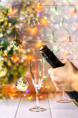 Men's hands open a bottle of champagne on the background of a New Year tree and garlands and glasses.