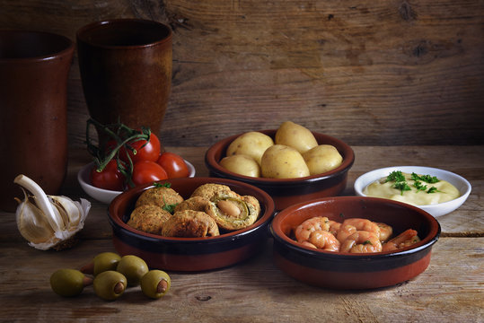 Rustic party appetizers such as baked olives, prawn shrimps, potatoes, tomato and garlic dip, spanish tapas served on a wooden board, dark background with copy space