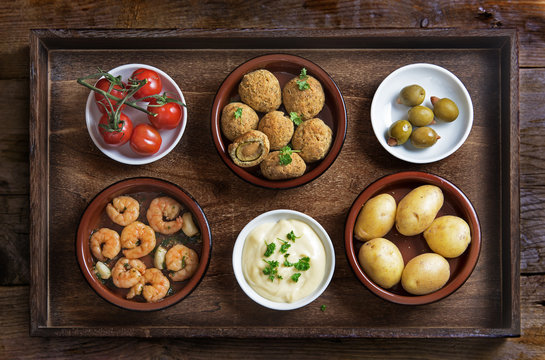 Spanish tapas such as baked olives, prawn shrimps, potatoes, tomato and garlic dip, party  appetizers on a rustic wooden tray, flat top view from above