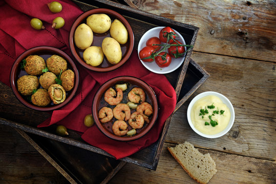 spanish tapas as party appetizers, baked olives, prawn shrimps, potatoes, tomato and garlic dip on a wooden tray with a red napkin on rustic wood, warm dark country style, flat top view from above