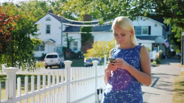 A woman is using a smartphone. It stands on the street near a white fence, behind the American flag. A typical American town
